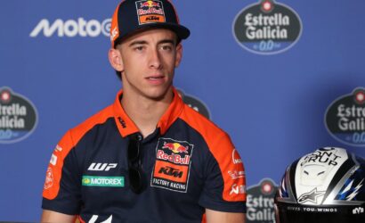 MotoGP rider in Red Bull KTM factory racing gear at a press conference, with helmet beside him against a blue backdrop.