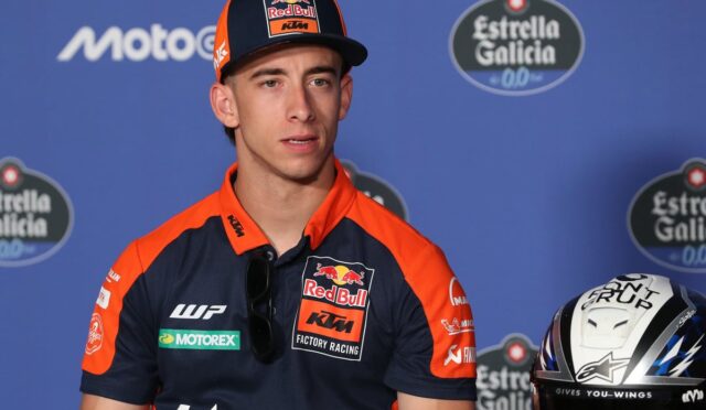 MotoGP rider in Red Bull KTM factory racing gear at a press conference, with helmet beside him against a blue backdrop.