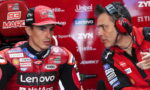 MotoGP rider in red Lenovo Ducati suit sits in a pit area, while a team engineer with red headphones talks to him against a backdrop of sponsor logos.