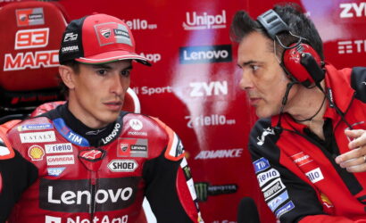 MotoGP rider in red Lenovo Ducati suit sits in a pit area, while a team engineer with red headphones talks to him against a backdrop of sponsor logos.