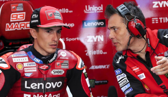 MotoGP rider in red Lenovo Ducati suit sits in a pit area, while a team engineer with red headphones talks to him against a backdrop of sponsor logos.