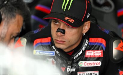 Close-up of a MotoGP rider in a Monster cap and racing suit with sponsor patches, focused in the pit area.
