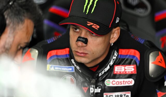 Close-up of a MotoGP rider in a Monster cap and racing suit with sponsor patches, focused in the pit area.