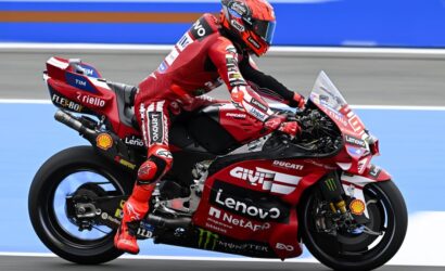 Professional motorcycle racer in red leathers on a red Ducati race bike, speeding on a blue racetrack adorned with sponsor logos.