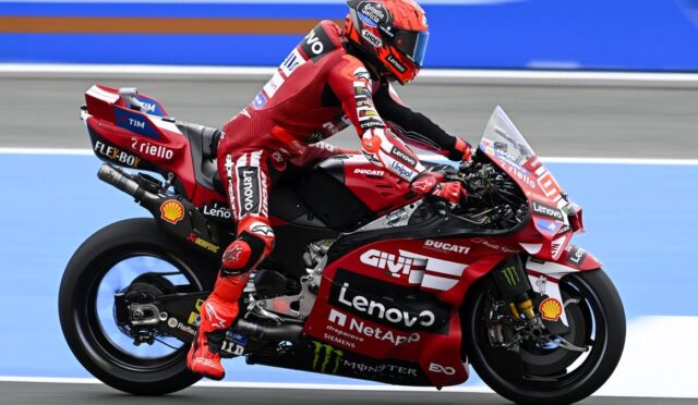 Professional motorcycle racer in red leathers on a red Ducati race bike, speeding on a blue racetrack adorned with sponsor logos.