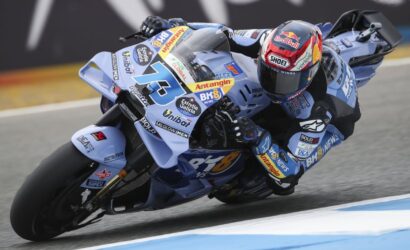 Racer on a light blue race bike leaning into a corner on a track, covered in sponsor stickers and a red‑bull helmet visor visible