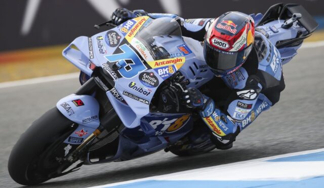 Racer on a light blue race bike leaning into a corner on a track, covered in sponsor stickers and a red‑bull helmet visor visible
