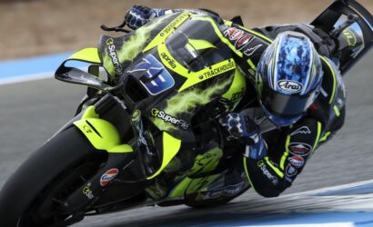 Motorcycle racer in a neon-yellow racing suit and bike leaning into a turn on a track, helmeted with blue visor and sponsor logos visible.