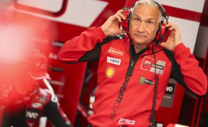 Pit crew member in a red Ducati team jacket wearing a headset, adjusting the ear cups in a red paddock.