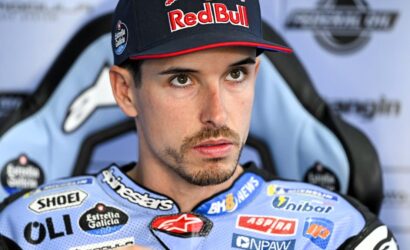 Racing driver wearing a Red Bull cap and blue sponsor-packed suit, seated in a race chair, looking pensive.