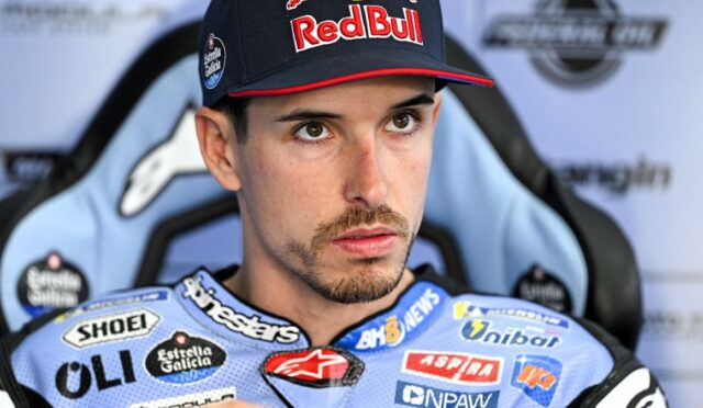 Racing driver wearing a Red Bull cap and blue sponsor-packed suit, seated in a race chair, looking pensive.