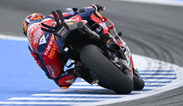 Moto racing rider in red-and-blue suit leans into a sharp turn on a blue-carpeted track, sponsor logos visible on gear and bike behind.
