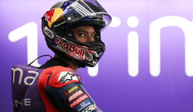 Close-up of a motorcycle racer in a Red Bull helmet, looking over the shoulder, with a purple backdrop behind.