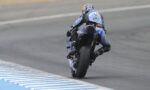 Motorcycle racer on a blue-and-black bike leaning into a turn on a race track, rear view