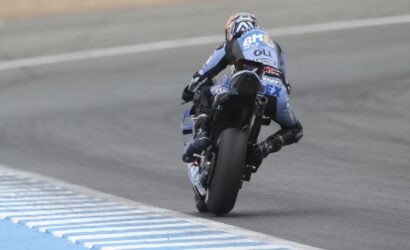 Motorcycle racer on a blue-and-black bike leaning into a turn on a race track, rear view