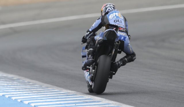 Motorcycle racer on a blue-and-black bike leaning into a turn on a race track, rear view