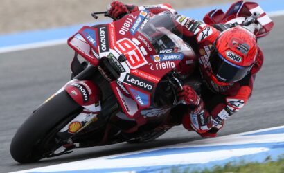 Motorcycle racer in red racing suit and helmet leaning into a left-hand turn on a track, bike covered in sponsor logos like Lenovo and Shell.