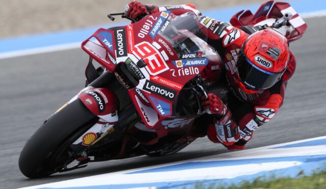 Motorcycle racer in red racing suit and helmet leaning into a left-hand turn on a track, bike covered in sponsor logos like Lenovo and Shell.