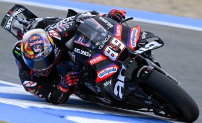 MotoGP rider in black/red gear leans into a sharp turn on a blue-marked race track, bike number 69.