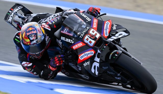 MotoGP rider in black/red gear leans into a sharp turn on a blue-marked race track, bike number 69.