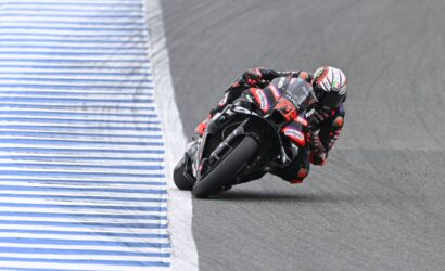 Motorcycle racer in a black-red suit leaning through a sharp turn on a race track beside a blue-and-white striped barrier.
