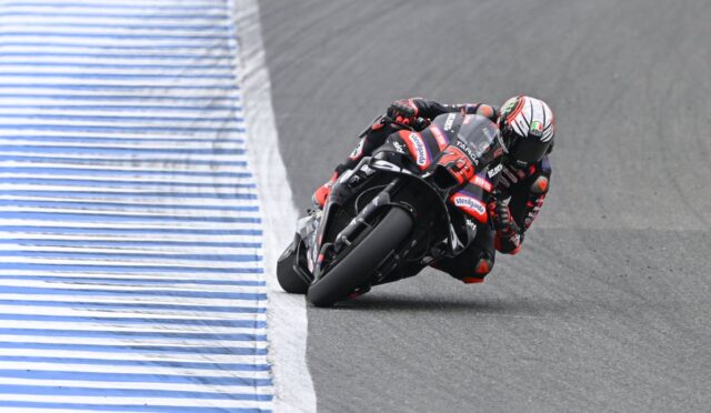 Motorcycle racer in a black-red suit leaning through a sharp turn on a race track beside a blue-and-white striped barrier.