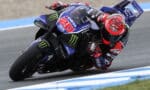 MotoGP rider in a red helmet leans into a turn on a blue Yamaha racing bike with sponsor logos on a race track.