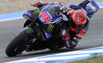 MotoGP rider in a red helmet leans into a turn on a blue Yamaha racing bike with sponsor logos on a race track.