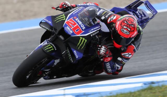 MotoGP rider in a red helmet leans into a turn on a blue Yamaha racing bike with sponsor logos on a race track.