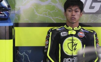 Motorcycle racer in neon-yellow and black racing suit stands against a lime backdrop with sponsor logos visible on the suit.