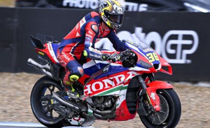 Motorcycle racer in red-blue Honda livery leaning into a turn on a race track, helmeted rider.