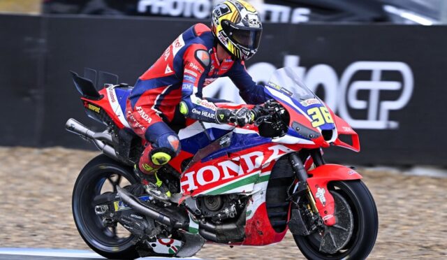 Motorcycle racer in red-blue Honda livery leaning into a turn on a race track, helmeted rider.