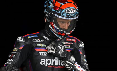 Professional motorcycle racer in an Aprilia Racing suit adjusts his gloves, wearing a colorful KYT helmet against a black background.