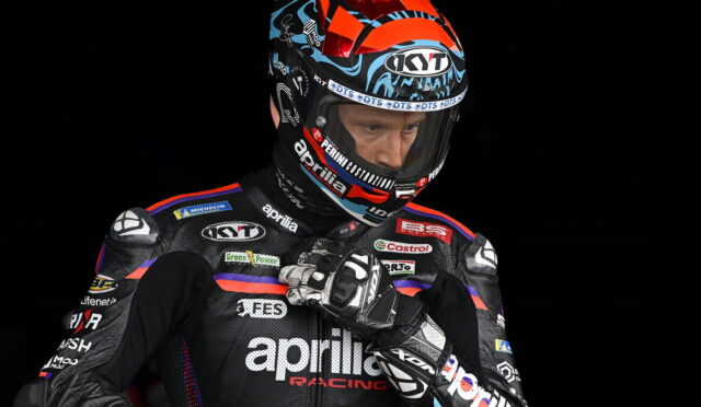 Professional motorcycle racer in an Aprilia Racing suit adjusts his gloves, wearing a colorful KYT helmet against a black background.