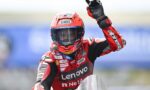 MotoGP rider in red Lenovo racing suit raises gloved hand on the track, helmet with Shoei logo.