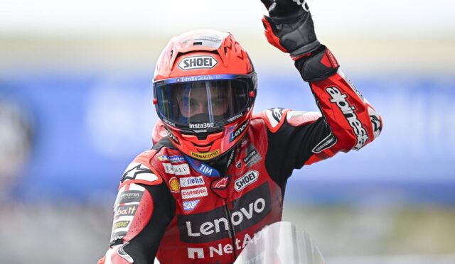MotoGP rider in red Lenovo racing suit raises gloved hand on the track, helmet with Shoei logo.