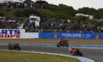 Motorcycle race on a curved track with three riders leaning into a turn as a large crowd watches from behind a fence and barriers with sponsor banners nearby.