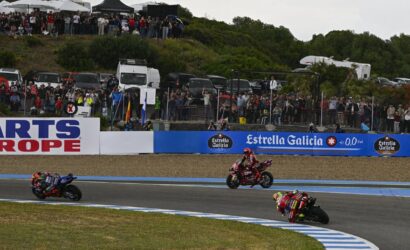 Motorcycle race on a curved track with three riders leaning into a turn as a large crowd watches from behind a fence and barriers with sponsor banners nearby.