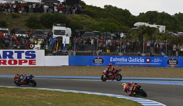 Motorcycle race on a curved track with three riders leaning into a turn as a large crowd watches from behind a fence and barriers with sponsor banners nearby.