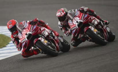 Three red-suited motorcycle racers lean into a track turn on shared bikes with sponsor decals visible on their bikes (MotoGP race)
