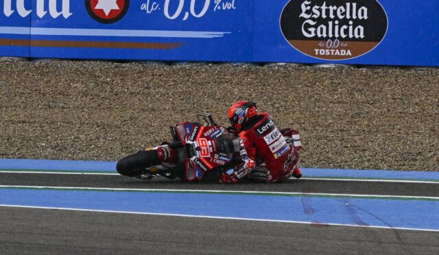 Two red‑leather MotoGP riders crash into a corner on a blue‑striped race track; gravel barrier behind and Estrella Galicia ad visible.