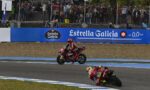 MotoGP race with two riders navigating a curve as spectators watch from behind a fence and blue banners show Estrella Galicia signage behind them.