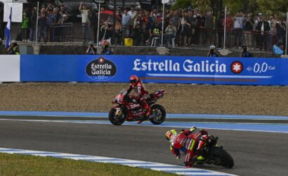 MotoGP race with two riders navigating a curve as spectators watch from behind a fence and blue banners show Estrella Galicia signage behind them.