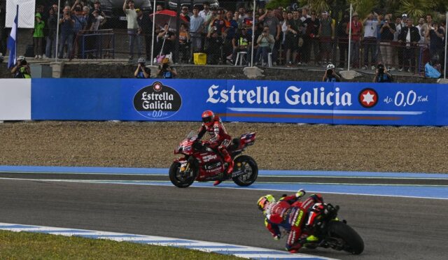 MotoGP race with two riders navigating a curve as spectators watch from behind a fence and blue banners show Estrella Galicia signage behind them.