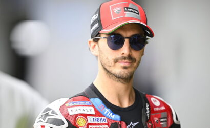 Motorcycle racer wearing a red cap and dark sunglasses, with a red sponsor-filled racing suit.