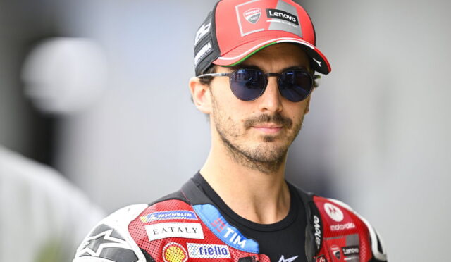 Motorcycle racer wearing a red cap and dark sunglasses, with a red sponsor-filled racing suit.