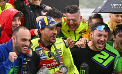 Group of male cyclists celebrating in the rain after a race, wearing neon racing suits and holding a medal and water bottles.
