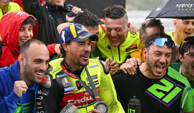 Group of male cyclists celebrating in the rain after a race, wearing neon racing suits and holding a medal and water bottles.