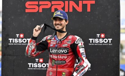 MotoGP rider in red Lenovo NetApp suit holds up a medal on the winner's podium with sponsor banners behind him (Sprint, Tissot).