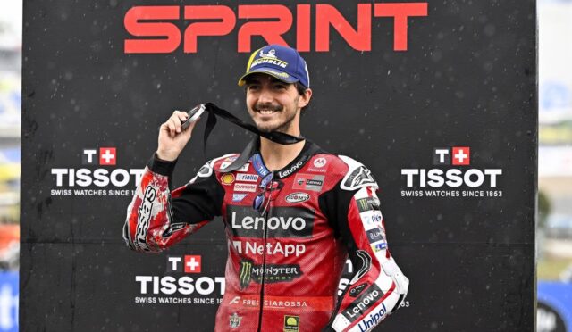 MotoGP rider in red Lenovo NetApp suit holds up a medal on the winner's podium with sponsor banners behind him (Sprint, Tissot).